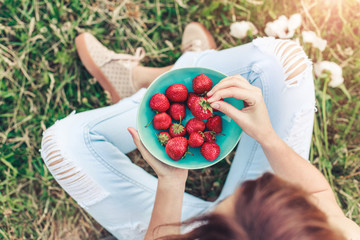 Girl in jeans sitting in summer grass and holding a plate of strawberries, knees and hands visible. Healthy breakfast, Clean eating, vegan food concept. Top view. Toning