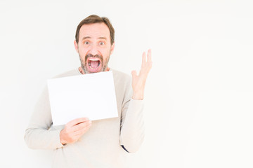 Senior man holding blank paper sheet over isolated background very happy and excited, winner expression celebrating victory screaming with big smile and raised hands