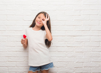 Young hispanic kid over white brick wall eating red heart lollipop candy with happy face smiling doing ok sign with hand on eye looking through fingers