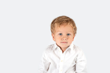 Half length portrait of a little serious boy wearing  white shirt.  He is siting on a white background in the studio