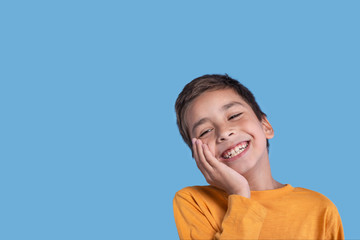 Close up emotional portrait of a boy wearing a yellow  on blue background in studio with copy space.He holds the cheek with his hand and laughs at something