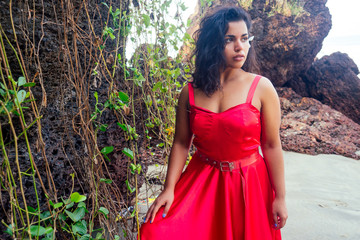 Curly-haired young afro american girl in a red long silk dress on a corset on the background of tropical rocks and vines.Female dark skin model indian woman enjoing at the sea coast paradise beach