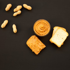 Bowl of peanut butter, toasts, unshelled peanuts on a black surface, top view. From above, overhead, flat lay.