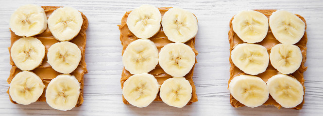 Vegan toasts with peanut butter and banana on a white wooden surface, overhead view.