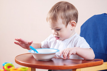 Little baby boy learning to eat at the kid table studying a plate and spoon in the kitchen
