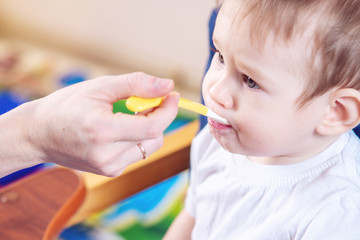 Little baby boy eating on a chair in the kitchen. Mom feeds holding in hand a spoon of porridge