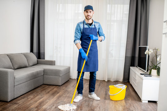 Portrait of a man as a professional cleaner in blue uniform washing floor with mopping stick and bucket in the living room of the apartment