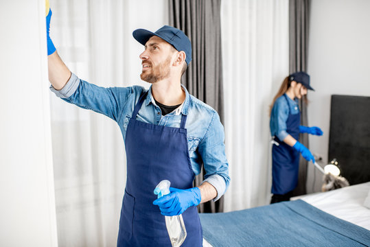 Couple As A Professional Cleaners In Uniform Rubbing Furniture And Wiping Dust In The Bedroom Or Hotel Room. Cleaning Service Concept