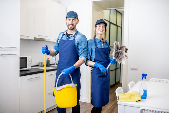 Portrait of a couple as a professional cleaners in uniform standing together with cleaning tools indoors