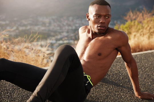 Healthy African American man relaxes on mountain road alone, being tired of morning training, poses outdoor, beautiful landscape in background. Handsome jogger works out for marathon in summer