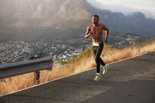 Athletic healthy man runs along road outdoors against mountain background, covers long distance, prepares for marathon. Sporty male exercises down hill, wears sportshoes, leggings, being in good shape
