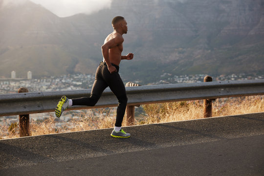 Sideways shot of athletic man covers long distance, runs on empty road, has early morning run on hilly path, workout at street, demonstrates his slim fit body, being strong. Determination concept