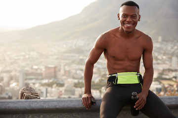 Sportsman with happy facial expression, shows white perfect teeth, wears leggings, holds bottle...