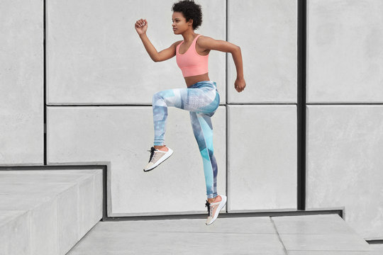 Sideways shot of athletic woman looks ahead, runs up stairs, wants to loose weight, has high jump, wears sportclothes, overcomes challenge, photographed in motion, burns fat in body. Exercising