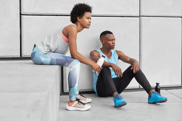 Fototapeta premium Photo of determined woman and man with dark skin, healthy body, have pensive contemplative facial expressions, relaxed Afro American girl sits at stairs near boyfriend, tired after playing basketball