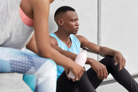 Cropped image of dark skinned male sits tired, his partner poses near with bottle of cold water, do sport together, lead active lifestyle, have regular trainings in gym or outdoor, wear sportsclothes