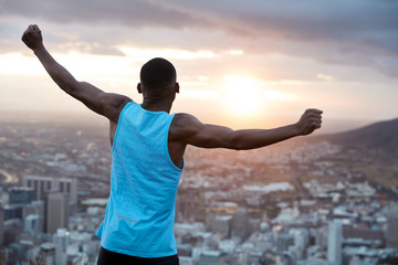 Fototapeta na wymiar Carefree independent man with dark skin, stands back at camera, stretches hands as holding world, feels freedom, wears blue casual vest, enjoys panoramic view with sunrise. Recreation concept