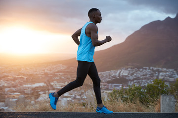 Outdoor view of active young male jogger covers long destiantion ealy in morning at dawn, runs over...
