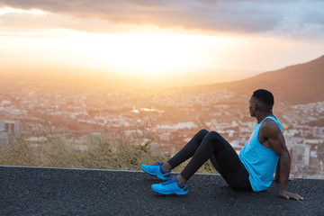 Horizontal view of restful athletic man with healthy body shape, muscles on arms, sits on high hill road, focused aside, admires majestic dawn, fresh air, has training outdoor. People, energy concept