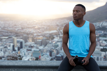 Fototapeta na wymiar Horizontal shot of handsome black man in casual outfit, holds bottle of water, being deep in thoughts, rests over city background with daylight, clear sky with copy space for your promotion.