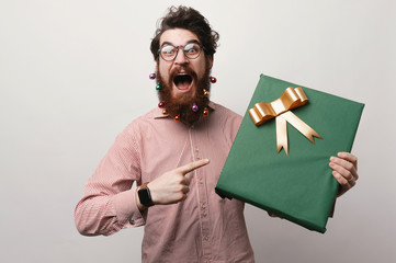 Happy and amazed man with decorated christmas beard pointing at big gift over white wall