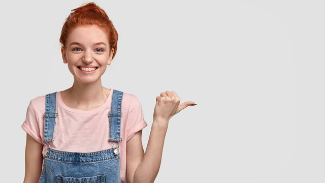 Studio shot of happy freckled young ginger girl with combed hair, has pleasant delighted facial expression, wears casual t shirt and dungarees, points with thumb aside, isolated over white background