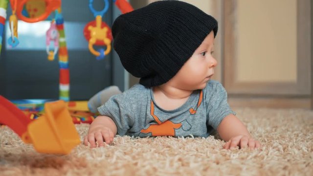 Adorable baby boy in a hat with a toys lying on the floor at home.