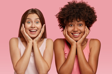 Glad beautiful multiethnic girls hold chins, recieve good news from friend, have broad smiles, model over pink studio wall, express pleasant emotions and feelings. Interracial relationship concept