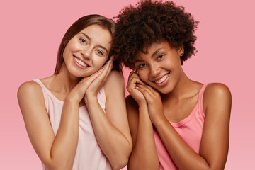 Pleased cheerful dark skinned young woman stands closely to her best Caucasian friend, model against pink background, express positive emotions and feelings. Multicultural companions pose indoor