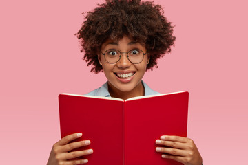 Horizontal shot of pretty smiling girl with pleasant facial expression, carries textbook, wears...