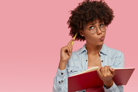 Image of contemplative dark skinned woman scratches head with pencil, pusrses lips and looks up with doubtful thoughtful expression, thinks on creative content as writes composition for class