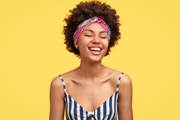 Joyful dark skinned model laughs pleasantly, closes eyes from happiness, recieves wonderful suggestion, being in high spirit during summer trip, wears headband and striped top, poses indoor.