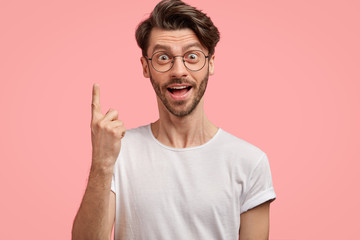 Surprised hipster with trendy haircut, has intriguing shocked gaze, points with index finger upwards, wears casual white t shirt and spectacles, isolated over pink background. Advertisement.