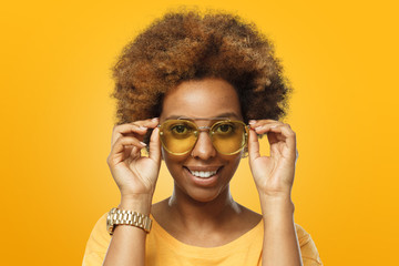 Young good-looking African female isolated on yellow background wearing trendy sunglasses and, holding rim of glasses with fingers, smiling happily