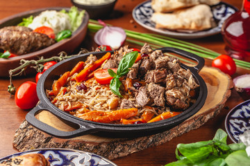 The concept of Uzbek, oriental cuisine. Pilaf in a cast-iron pan, with pieces of lamb lies on a wooden table. Festive table set.