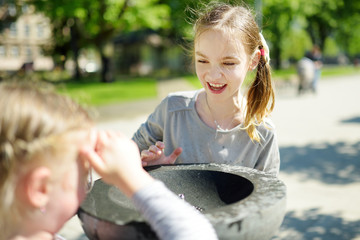 Two sisters having fun with drinking water fountain on warm and sunny summer day.