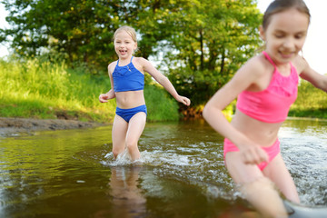 Two little sisters having fun on a sandy lake beach on warm and sunny summer day. Kids playing by the river.