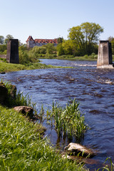 Natural landscape with river and ancient castle; At the forefront there are ancient bridge supports, large stones on the embankment; On the back of the background is a bright castle with red roof tile