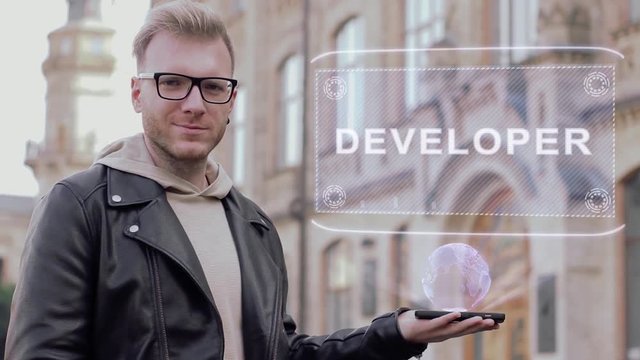 Smart young man with glasses shows a conceptual hologram Developer. Student in casual clothes with future technology mobile screen on university background