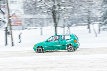 Driving in snow. Motion in blur small green car in heavy snowfall in city road. Abstract blur winter weather background