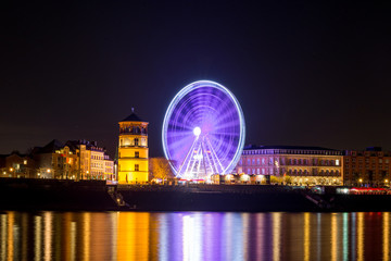 Obraz premium Night scenery colorful illuminated and reflected on river of buildings in old town, big moving Ferris Wheel and Christmas Market on promenade along riverside Rhine River in Düsseldorf, Germany. 