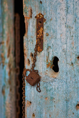 Rusting vintage iron padlock and chain hanging from a derelict wooden blue painted door on an abandoned farm house in rural Greece.