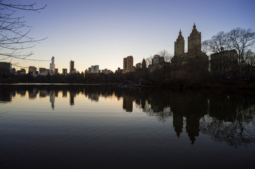 Tranquil sunset view of the scenic skyline silhouette of the Upper West Side reflecting on the flat surface of the Central Park Lake on a calm winter evening in New York City