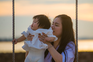 young sweet and happy Asian Chinese woman holding baby girl swinging together at beach swing on Summer sunset in mother and little daughter love concept