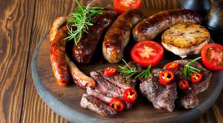 Close-up photo of mixed grilled meat platter. Beef, pork, poultry, sausages, grilled garlic, chili pepper, red tomatoes on wooden rustic background.
