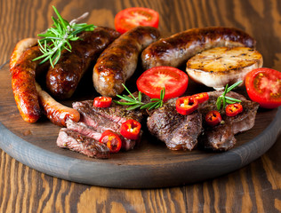 Close-up photo of mixed grilled meat platter. Beef, pork, poultry, sausages, grilled garlic, chili pepper, red tomatoes on wooden rustic background.