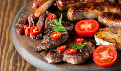 Close-up photo of mixed grilled meat platter. Beef, pork, poultry, sausages, grilled garlic, chili pepper, red tomatoes on wooden rustic background.
