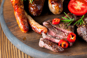 Close-up photo of mixed grilled meat platter. Beef, pork, poultry, sausages, grilled garlic, chili pepper, red tomatoes on wooden rustic background.