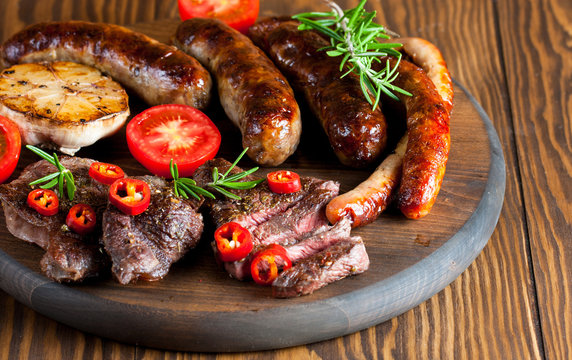 Close-up photo of mixed grilled meat platter. Beef, pork, poultry, sausages, grilled garlic, chili pepper, red tomatoes on wooden rustic background.
