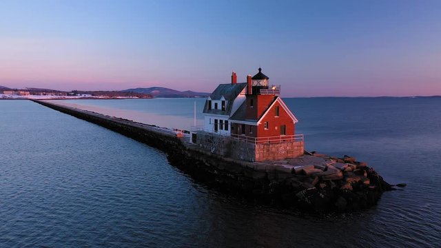 Low aerial ORBIT around a red brick lighthouse at the end of a snow dusted rocky breakwater revealing a setting full moon